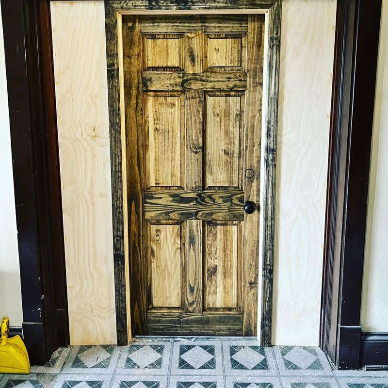 Rustic six-panel wood door in a dark wood frame with plywood filler panels beside it