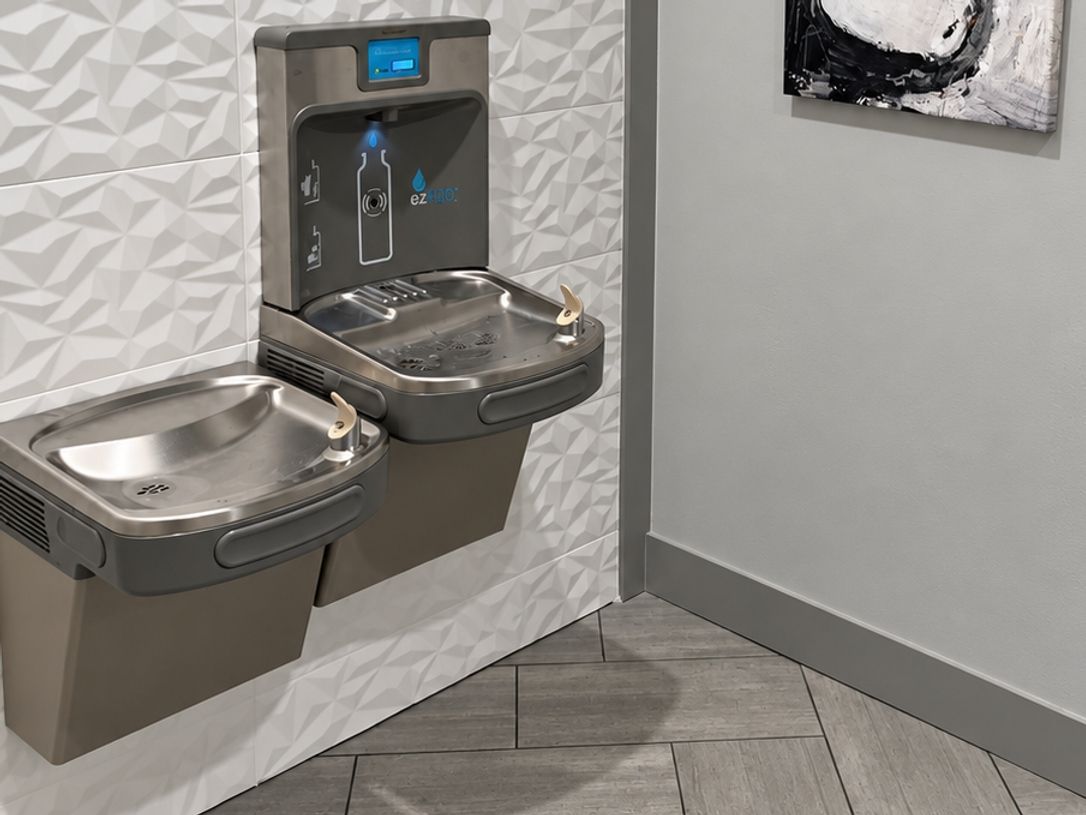 White three-dimensional tile wall with a stainless drinking fountain and bottle filler above herringbone gray floor tile