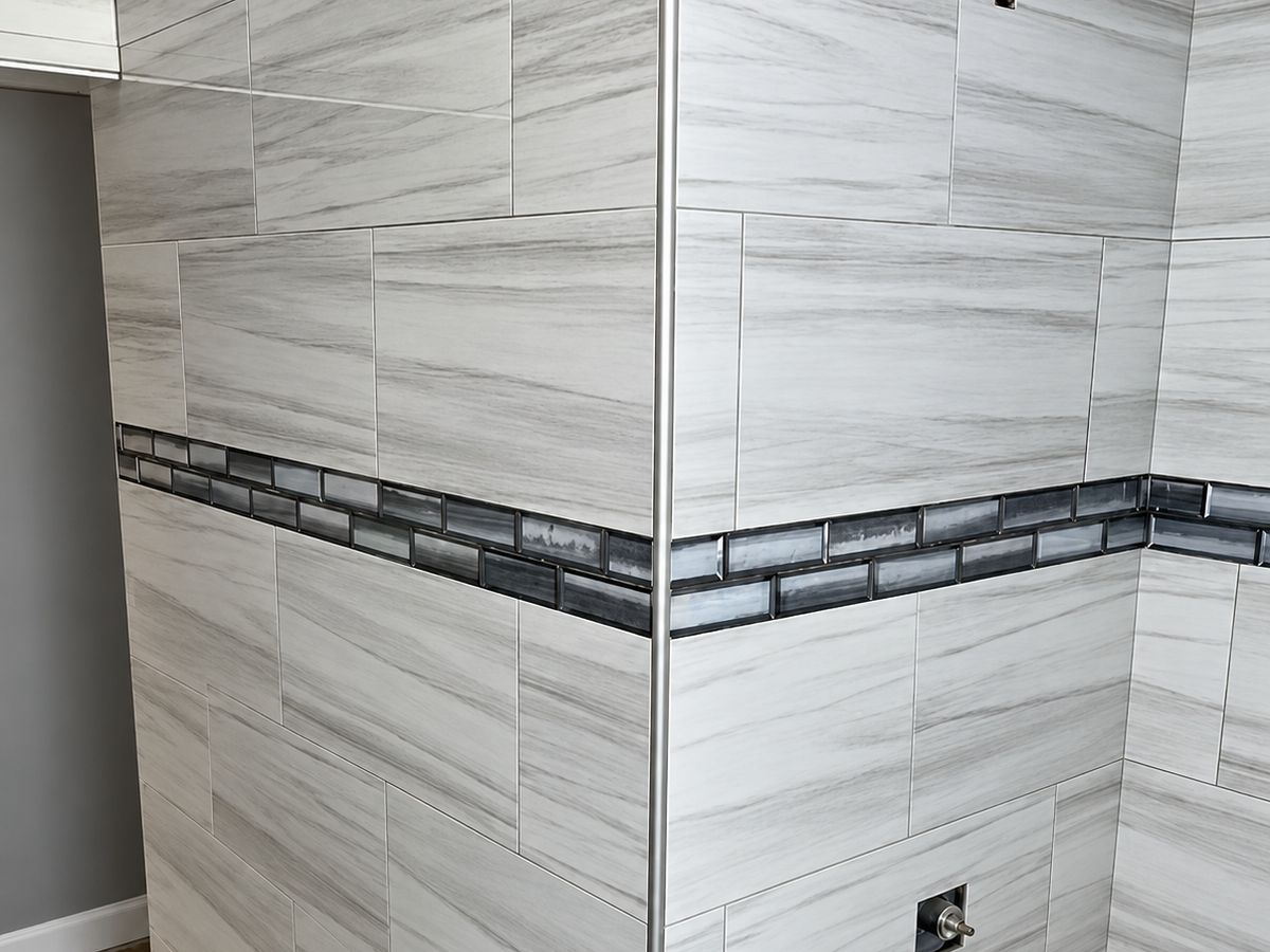 Corner of a bathroom tub surround tiled in large white and gray wood-grain porcelain with a band of gray glass subway tile