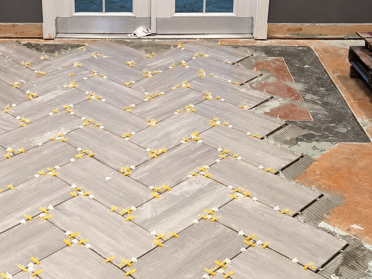 Gray wood-look plank porcelain tiles being laid in a herringbone pattern on a concrete floor