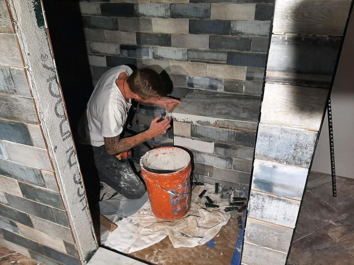 Tile setter in a white t-shirt kneeling to set blue-gray and white stained-wood-look rectangular tiles inside a shower
