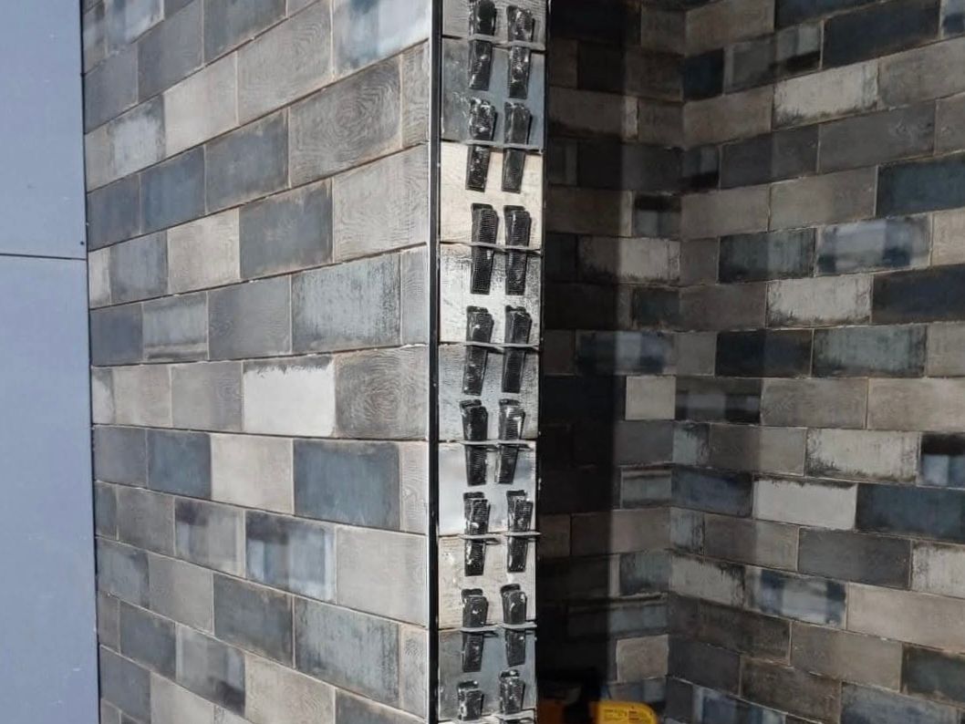 Corner of a shower area tiled in blue, gray, and cream wood-look tile with clips and a metal edge strip