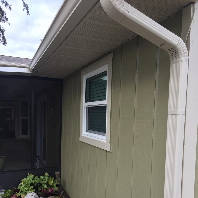 White-framed window in green siding beside a downspout above a flower bed